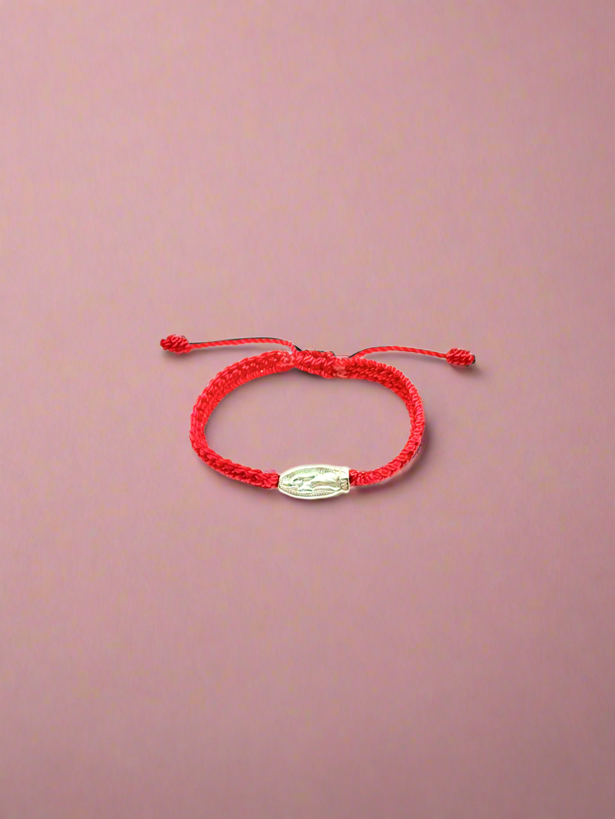 Front view of adjustable red braided baby bracelet with silver Our Lady of Guadalupe charm and silver beads, fits infants 0–12 months on a pink background