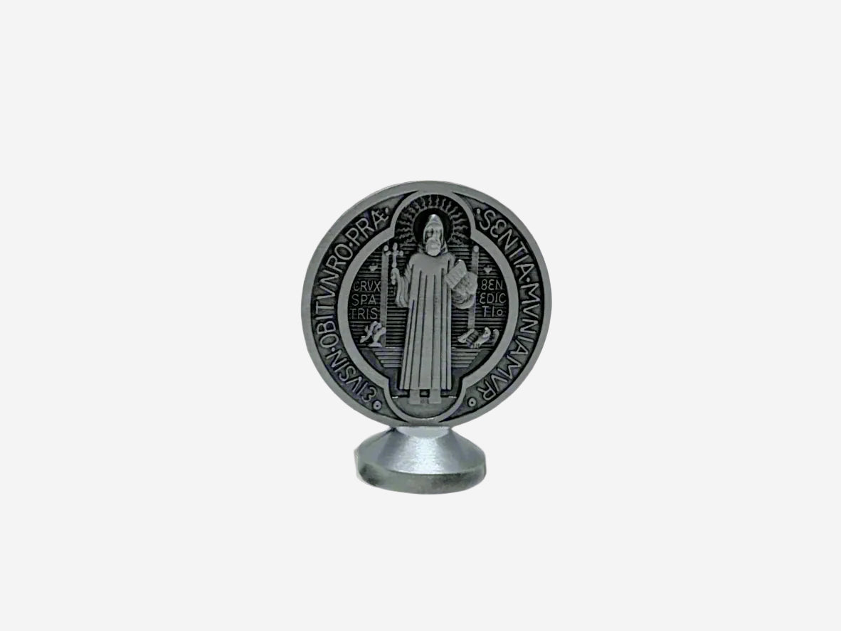 A back view Dashboard St Benedict medal in silver displayed upright on a small stand. The front of the medal features traditional Latin inscriptions and symbols associated with St. Benedict, including the cross and initials representing prayers of protection. The background is plain and neutral, highlighting the details of the medal.