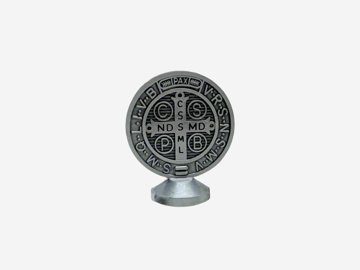 A Dashboard St Benedict medal in silver displayed upright on a small stand. The front of the medal features traditional Latin inscriptions and symbols associated with St. Benedict, including the cross and initials representing prayers of protection. The background is plain and neutral, highlighting the details of the medal.