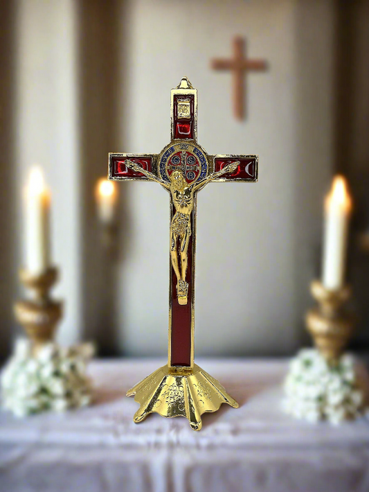 Decorative St. Benedictcrucifix with gold and red accents on a table with candles and flowers.
