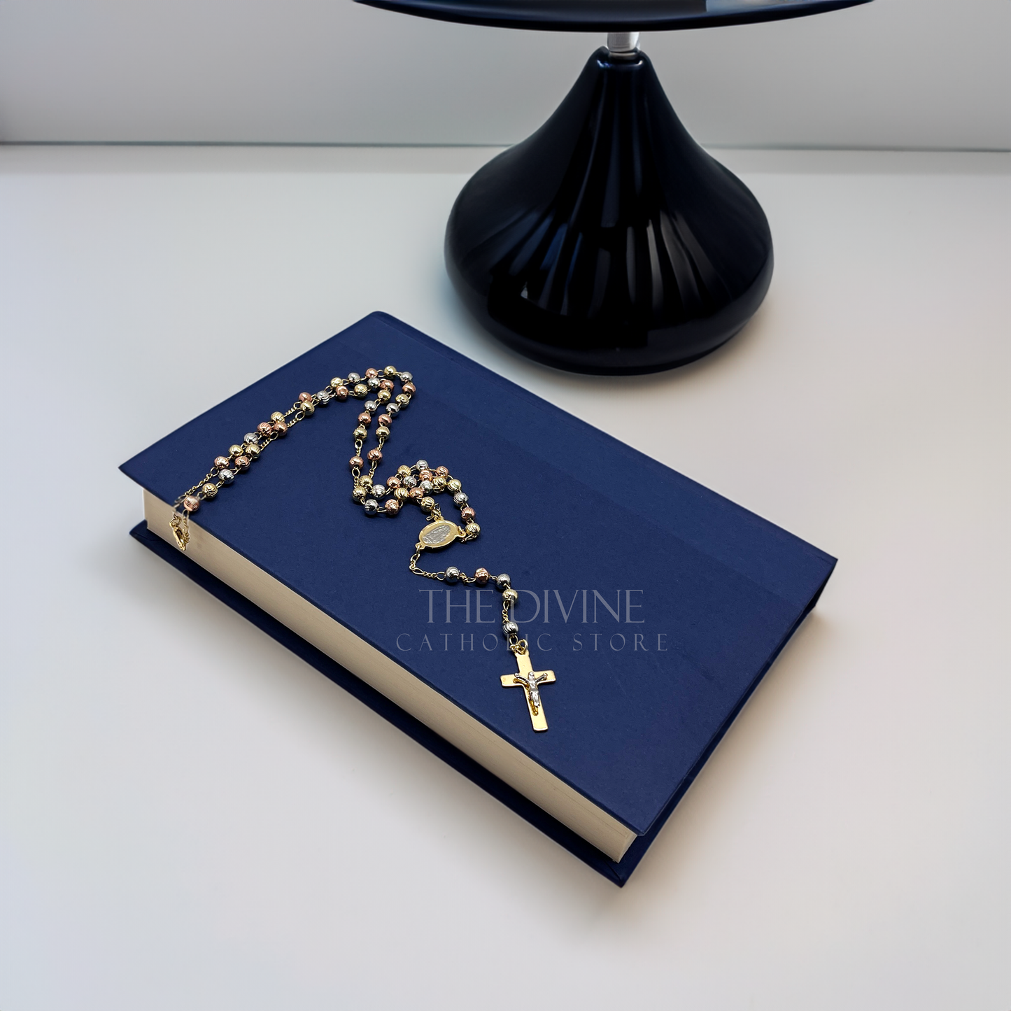 Elegant 18k gold plated rosary prayer beads with Our Lady of Guadalupe centerpiece sitting on a blue book and white background next to a black lamp.