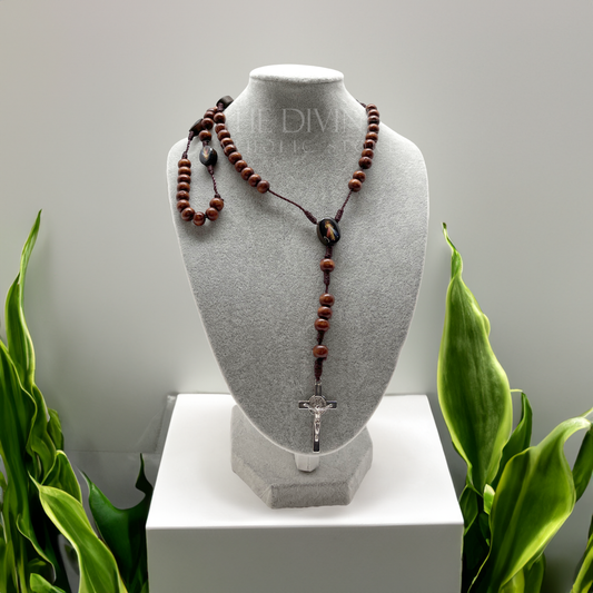 Divine Mercy wooden rosary displayed on gray jewelry bust, 17-inch Catholic prayer beads with St. Benedict crucifix on a white table top and white background surrounded by green plants.