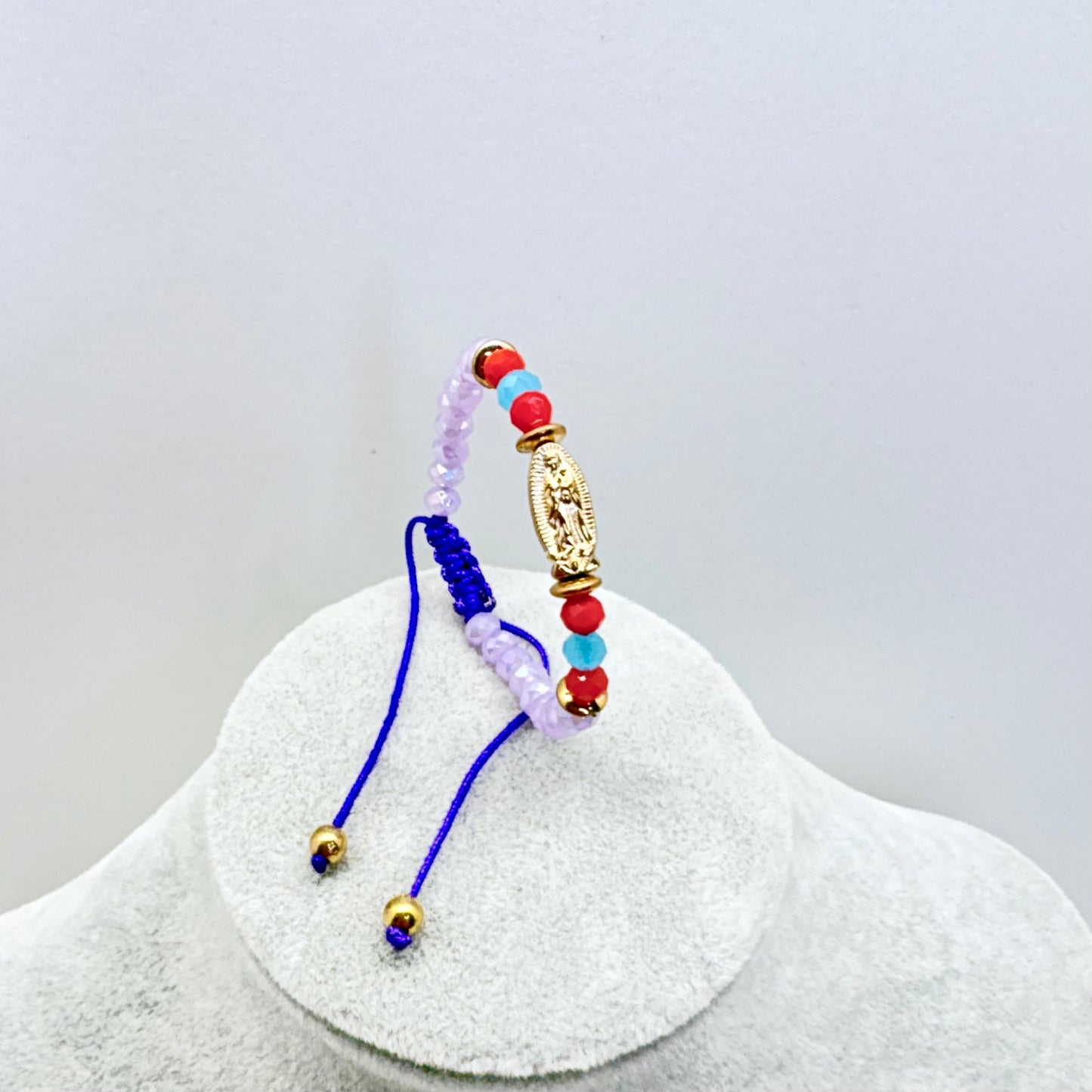 Beaded baby bracelet with Our Lady of Guadalupe medal shown upright on stand with purple beads and adjustable cord detail
