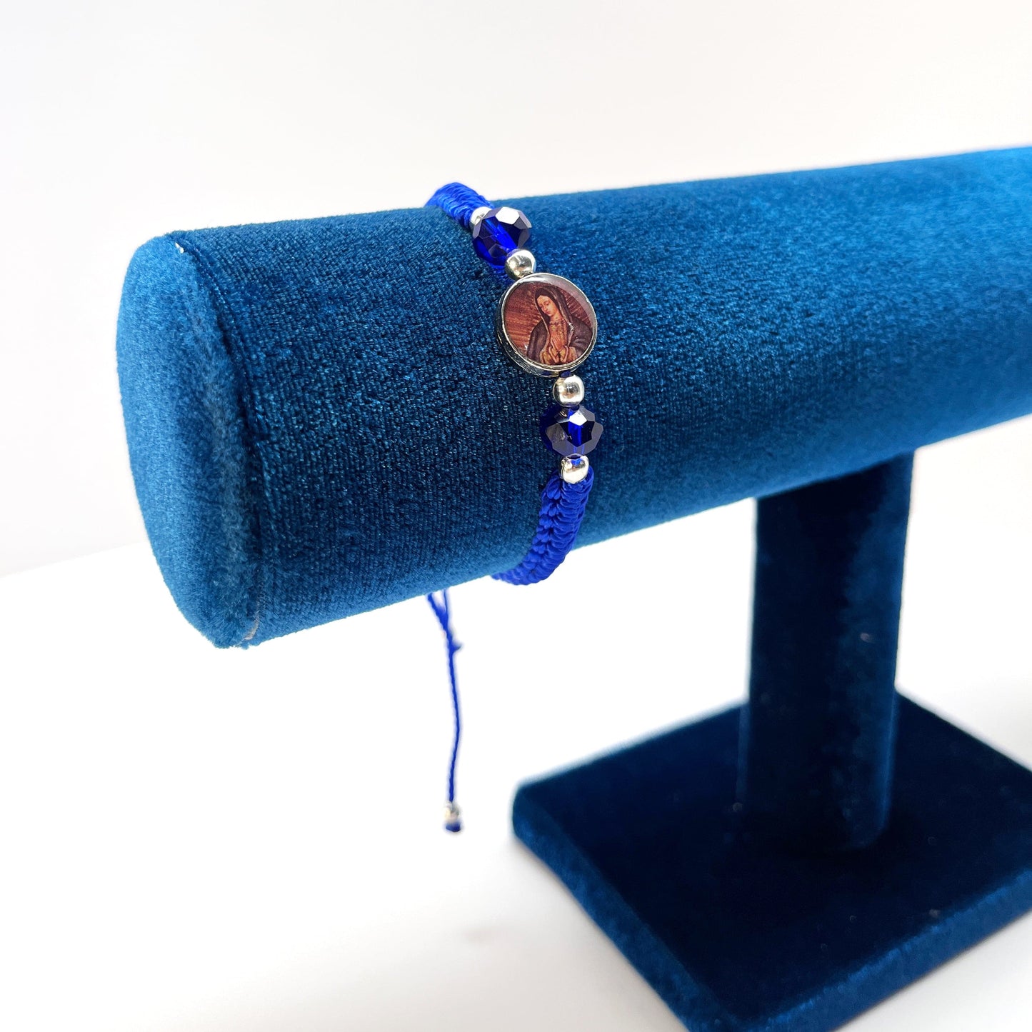 Upright view of a adjustable Catholic bracelet in blue nylon thread featuring rhinestones, silver beads, and Guadalupe charm on a blue bracelet holder.