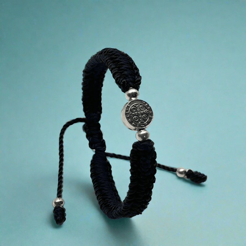 Upright view of a adjustable black nylon St. Benedict bracelet with woven cord and silver medal, durable and lightweight on a blue background.
