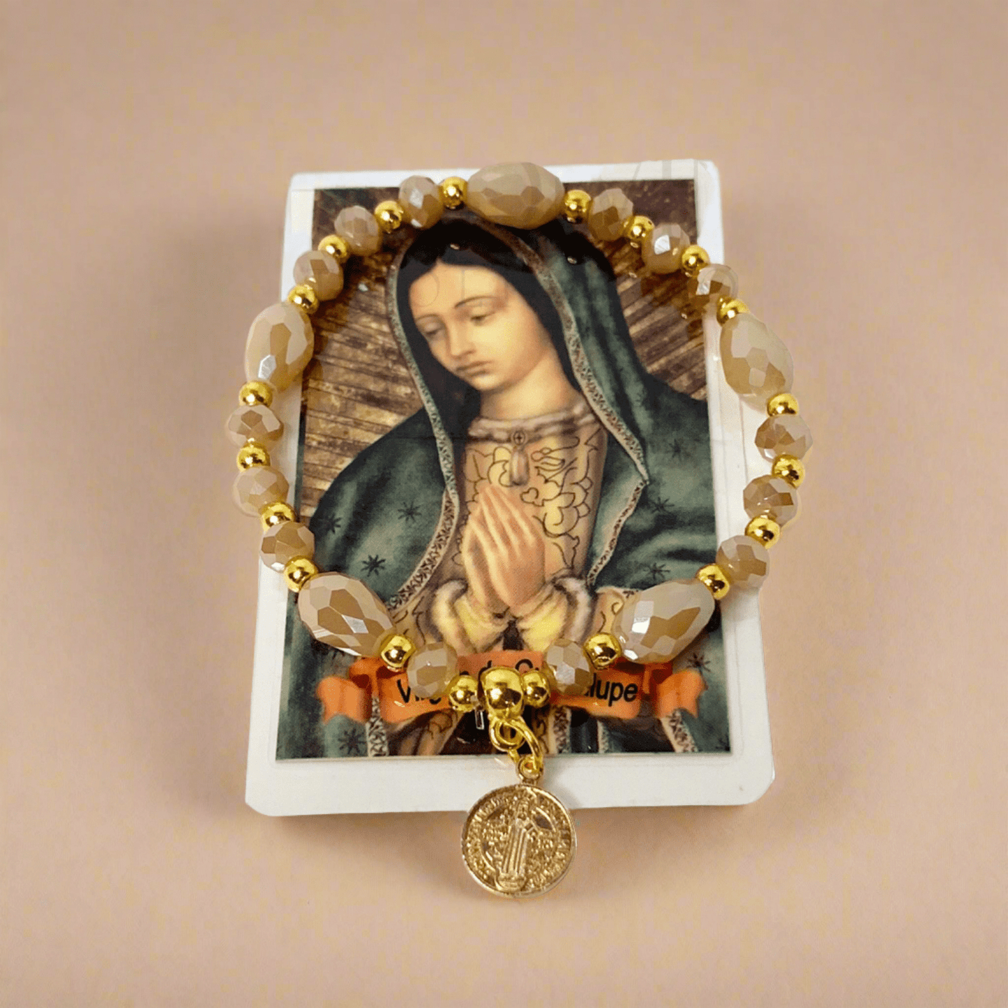 Beige and gold beaded St. Benedict bracelet with elastic band, shown with Spanish Our Lady of Guadalupe prayer card on a light brown background.
