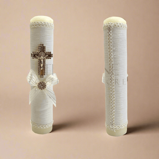 Single ivory baptism candle, decorated with ivory fabric, embroidered with ivory thread, gold colored St. Benedict crucifix outlined in rhinestones, and ivory bow with rhinestone in middle, showing front and back view of candle, with orange background.