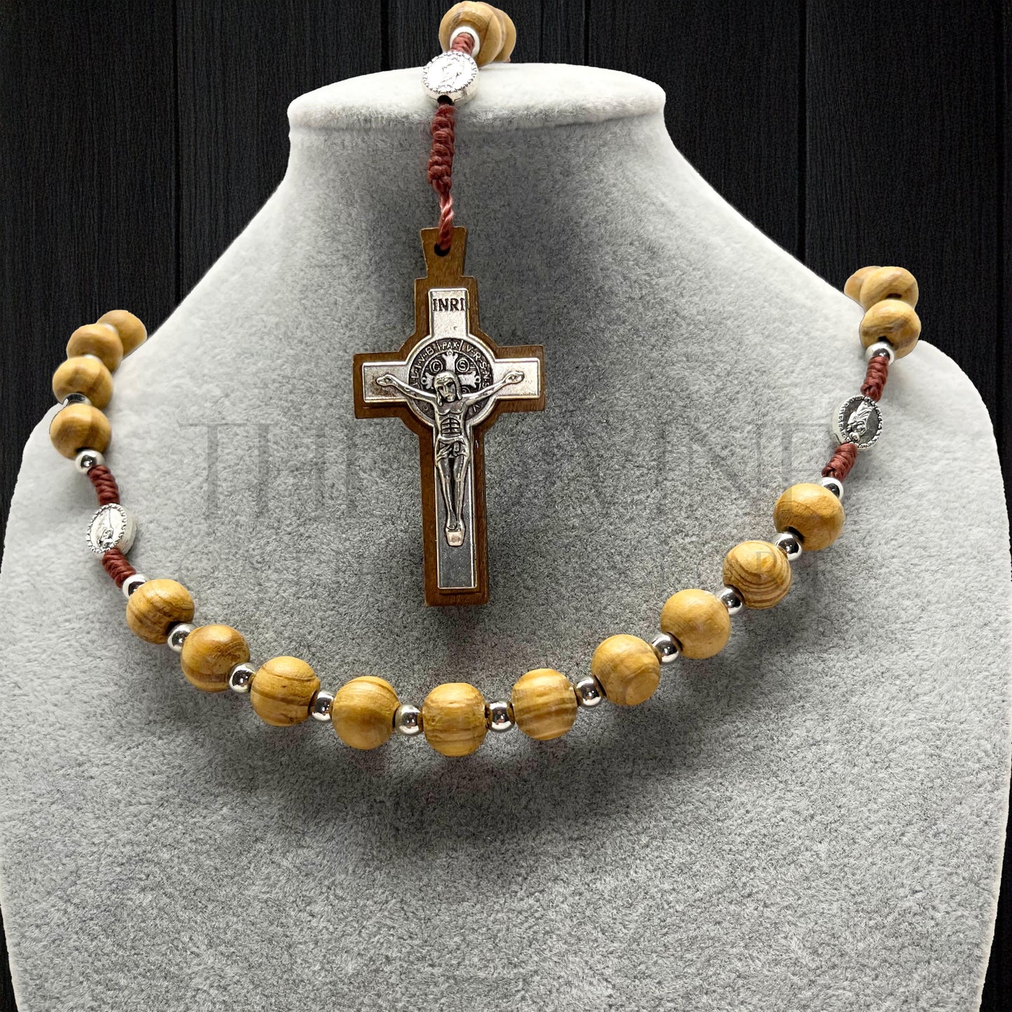 Beige wooden rosary beads with St. Jude and St. Benedict centerpiece and a close up of the silver St. Benedict crucifix displayed on gray bust against black backdrop