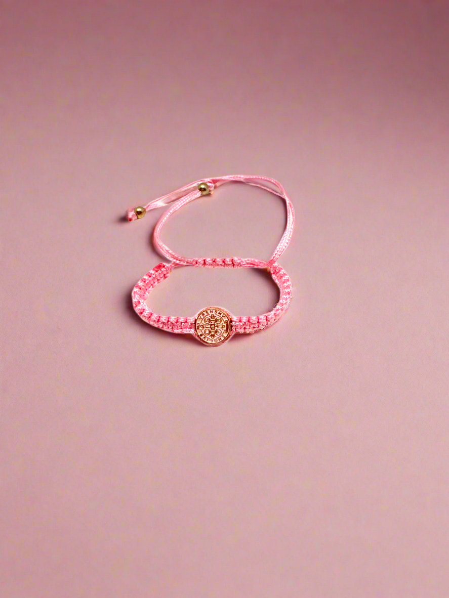 Pink St. Benedict baby bracelet braided bracelet with a circular St. Benedict medal charm on a pink background.