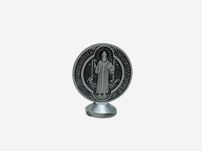  A back view Dashboard St Benedict medal in silver displayed upright on a small stand. The front of the medal features traditional Latin inscriptions and symbols associated with St. Benedict, including the cross and initials representing prayers of protection. The background is plain and neutral, highlighting the details of the medal.