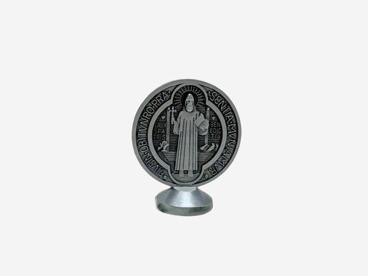  A back view Dashboard St Benedict medal in silver displayed upright on a small stand. The front of the medal features traditional Latin inscriptions and symbols associated with St. Benedict, including the cross and initials representing prayers of protection. The background is plain and neutral, highlighting the details of the medal.
