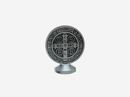  A Dashboard St Benedict medal in silver displayed upright on a small stand. The front of the medal features traditional Latin inscriptions and symbols associated with St. Benedict, including the cross and initials representing prayers of protection. The background is plain and neutral, highlighting the details of the medal.