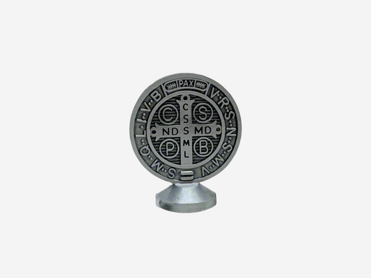  A Dashboard St Benedict medal in silver displayed upright on a small stand. The front of the medal features traditional Latin inscriptions and symbols associated with St. Benedict, including the cross and initials representing prayers of protection. The background is plain and neutral, highlighting the details of the medal.