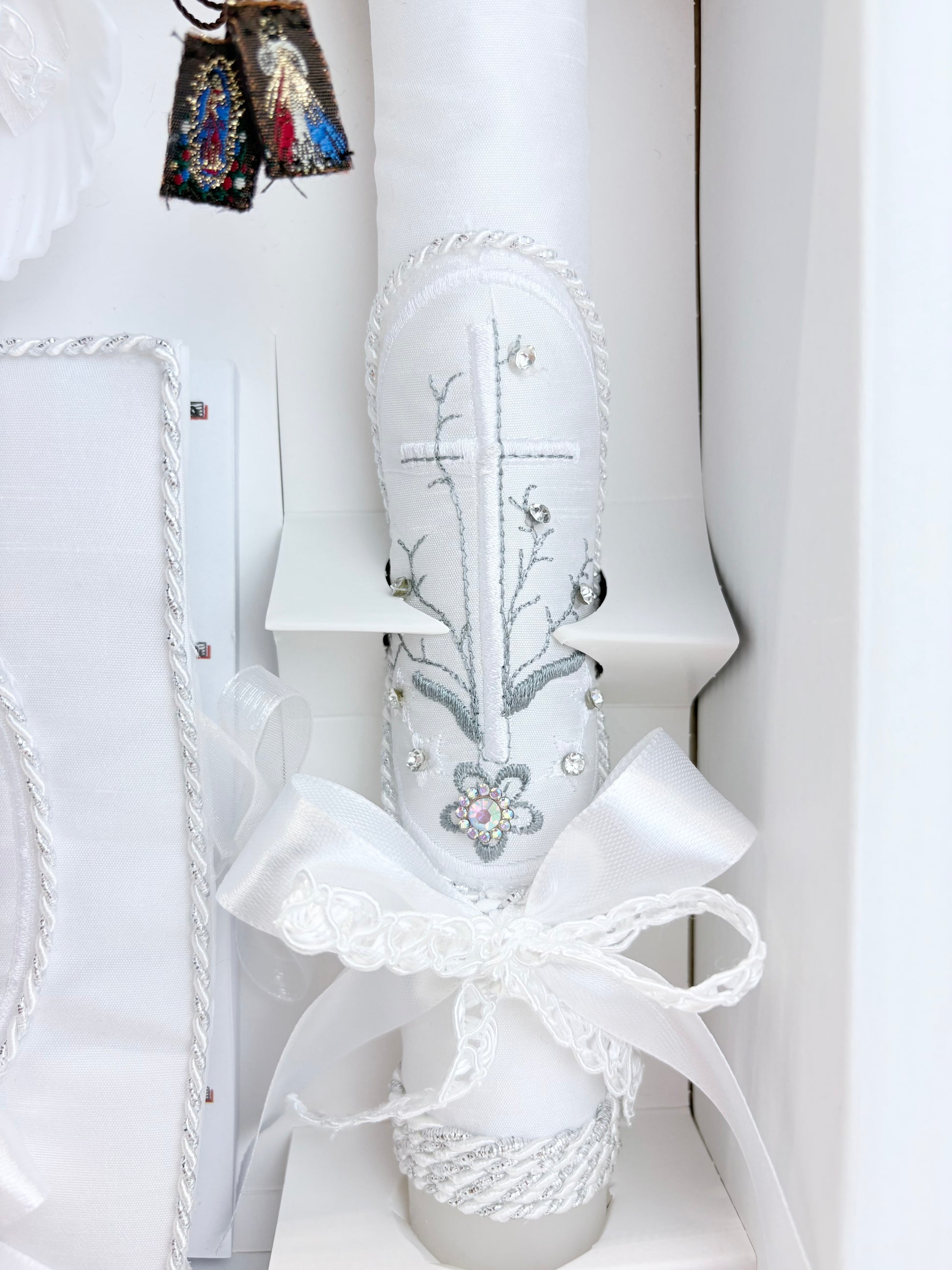 Closeup of white baptism candle embroidered cross with floral branch rhinestone accents and divine mercy guadalupe scapular
