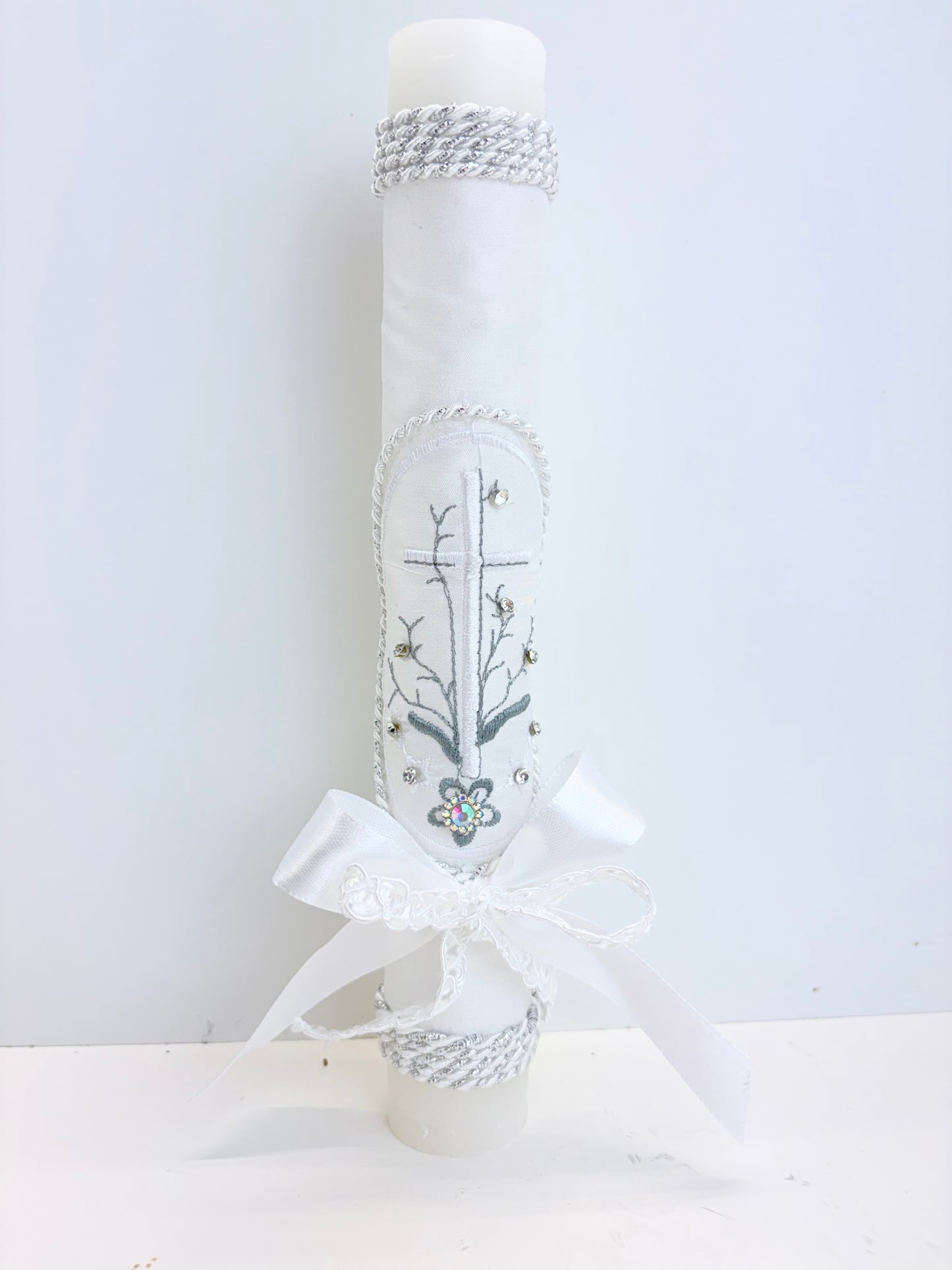 Front view of white baptism candle with embroidered cross floral branch design rhinestone accents silver rope trim and satin bow