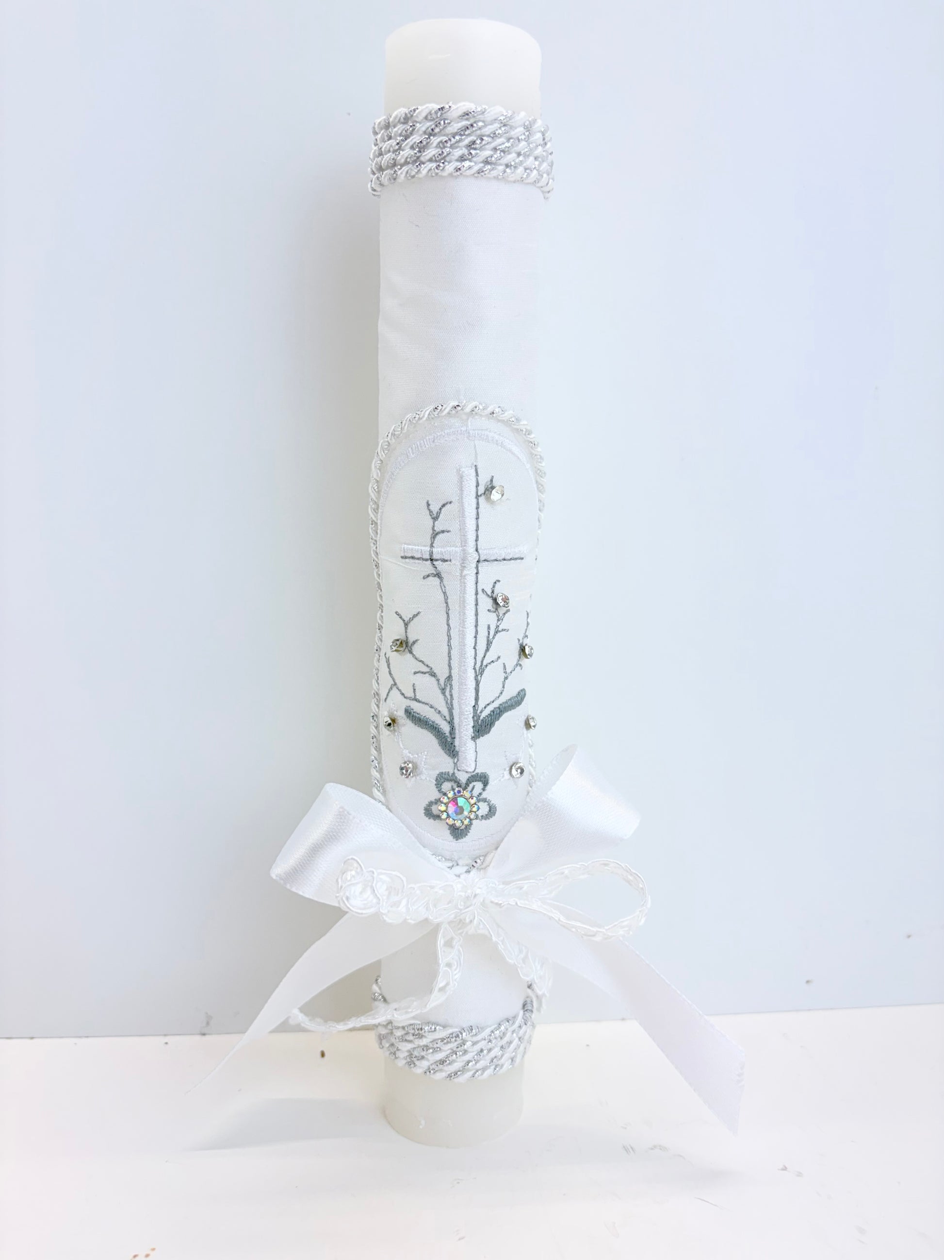 Front view of white baptism candle with embroidered cross floral branch design rhinestone accents silver rope trim and satin bow