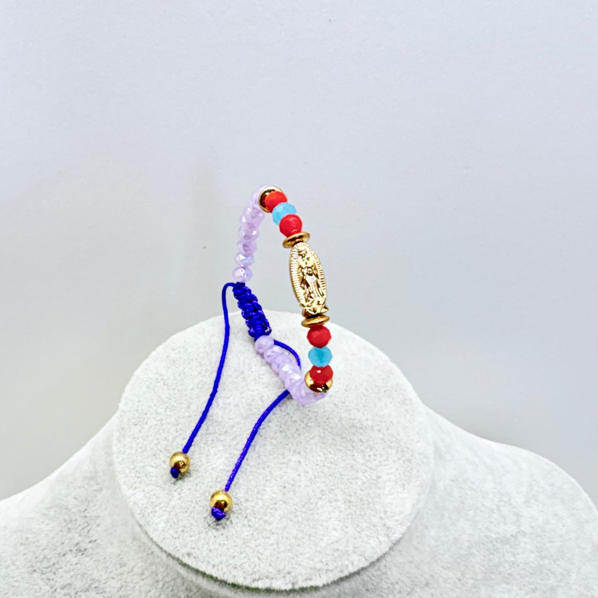 Beaded baby bracelet with Our Lady of Guadalupe medal shown upright on stand with purple beads and adjustable cord detail