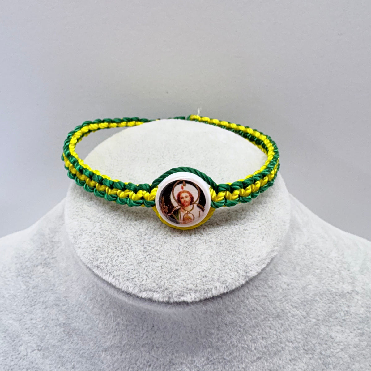 Green and yellow St. Jude bracelet displayed on gray jewelry stand, adjustable nylon Catholic bracelet with round saint charm