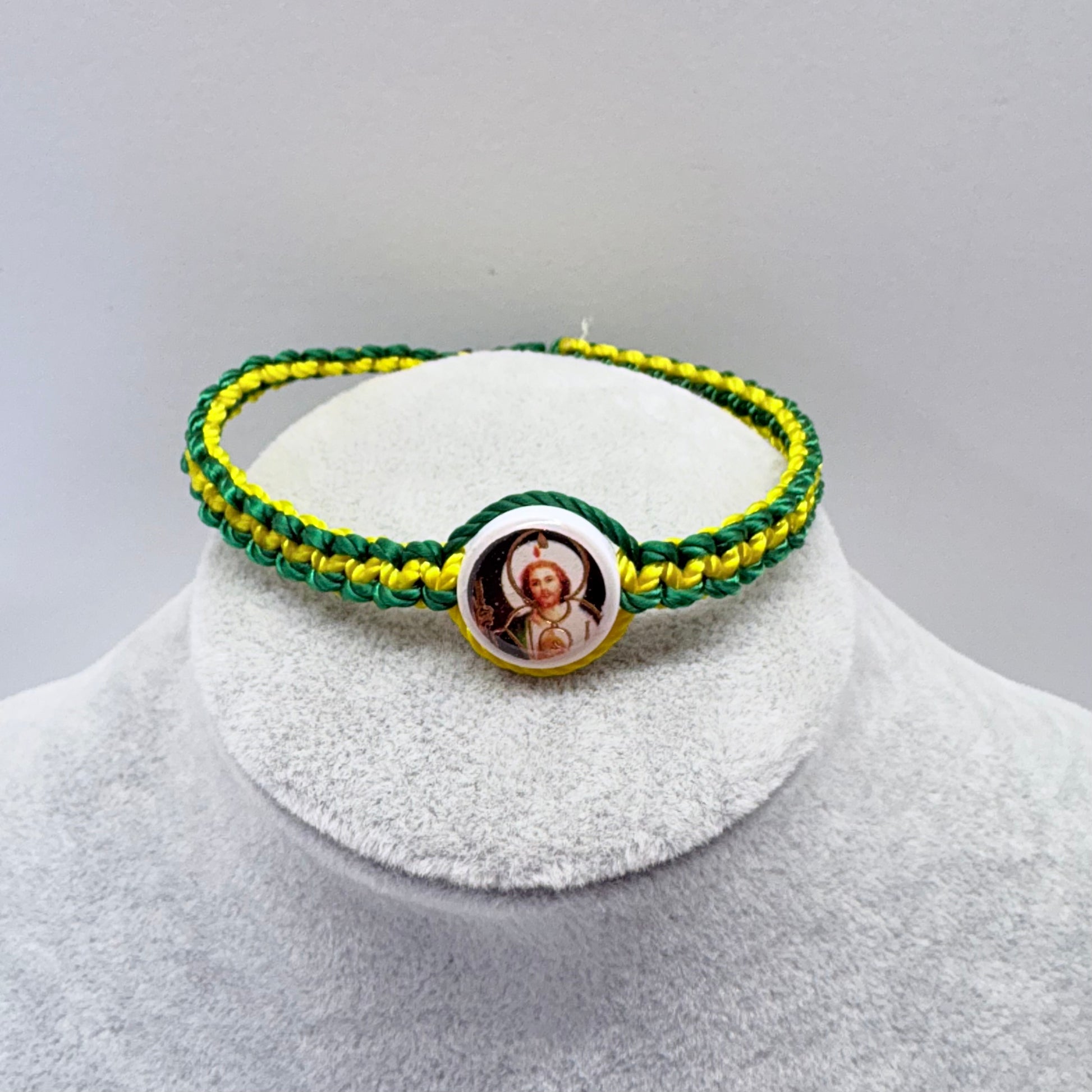 Green and yellow St. Jude bracelet displayed on gray jewelry stand, adjustable nylon Catholic bracelet with round saint charm