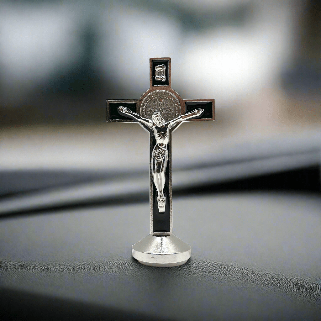 This 3 inch dashboard St. Benedict crucifix is black with a silver St.Benedict medal behind a silver corpus, silver-tone detailing and sturdy base with adhesive at bottom, sitting on a car dashboard.