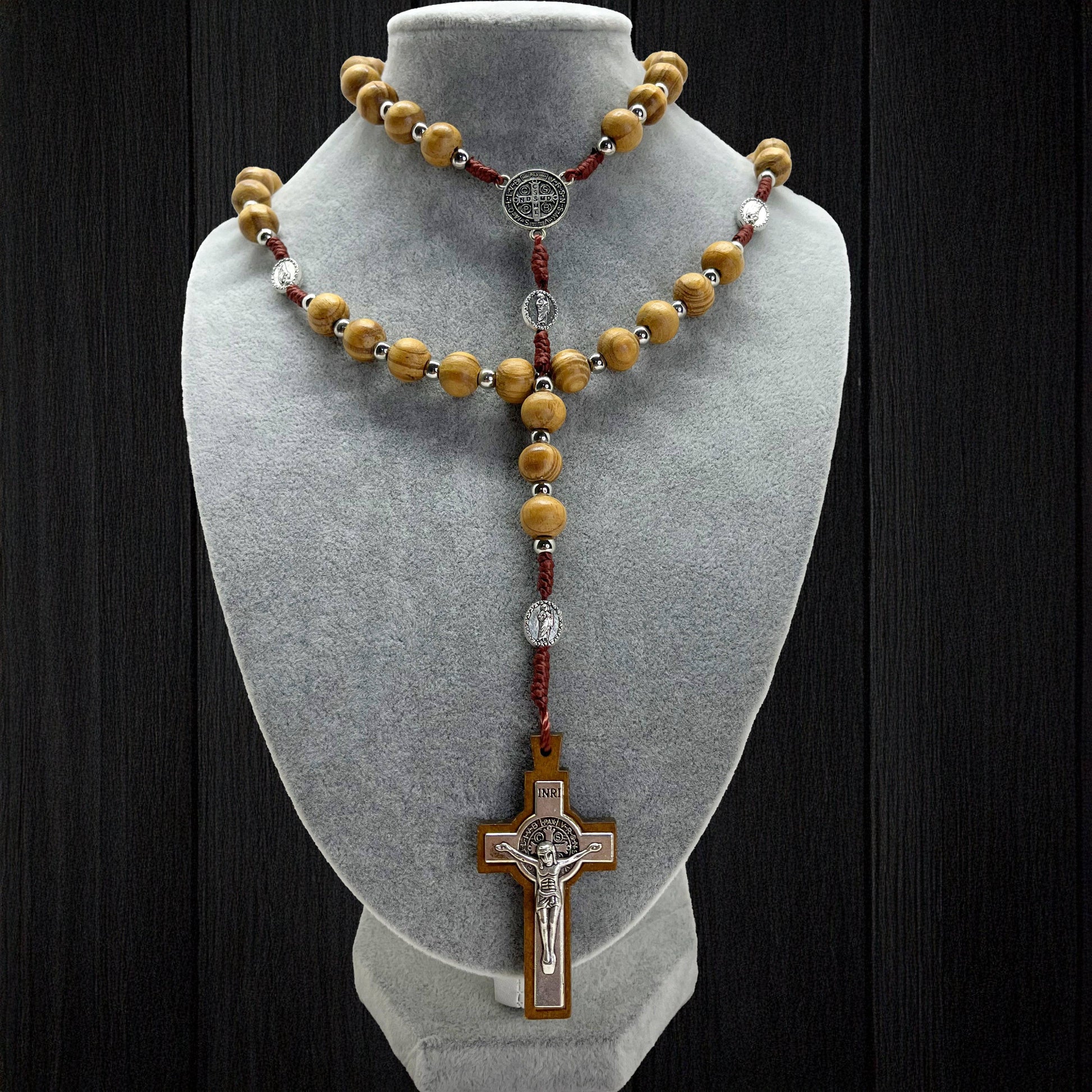 Handmade beige wooden rosary with St. Jude and St. Benedict medals displayed on gray jewelry bust against black background.