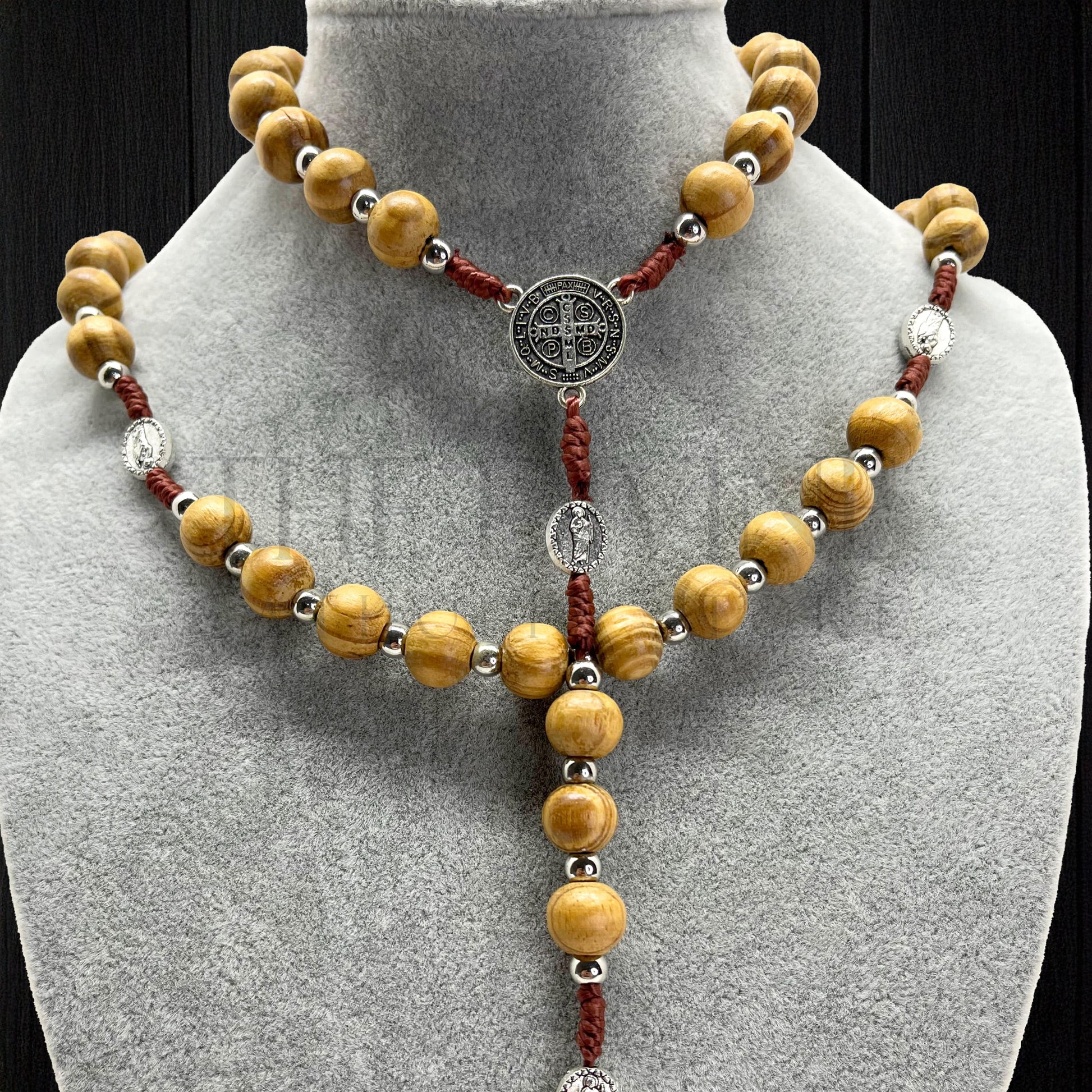 Catholic wooden rosary with beige beads and St. Jude and St. Benedict medals displayed on gray jewelry bust, black background.